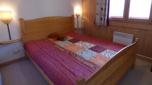Un lit en bois recouvert d’une courtepointe à motifs, deux lampes fixées au mur, une fen être avec des rideaux et un radiateur.