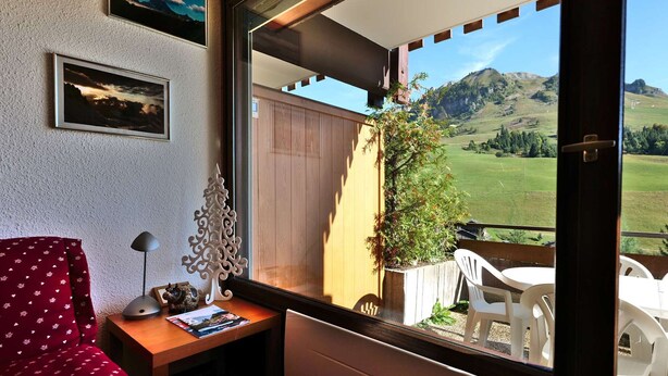 A room with a wooden desk, a red chair, a white radiator, and a view of a green landscape through a large window. - Comfortable 40m² apartment for 6 near ski slopes, large terrace (Le Grand-Bornand)