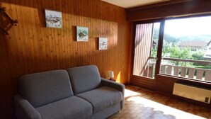 Una habitación acogedora con un sofá gris, una pequeña mesa redonda, paredes de madera y un amplio ventanal con vistas al balcón.