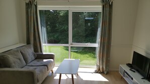 A living room with a sofa, a small white table, a TV on a stand, and a large window with curtains.