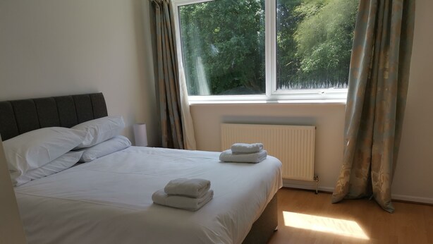 A neatly made bed with white linens and two folded towels, a window with curtains, and a radiator.