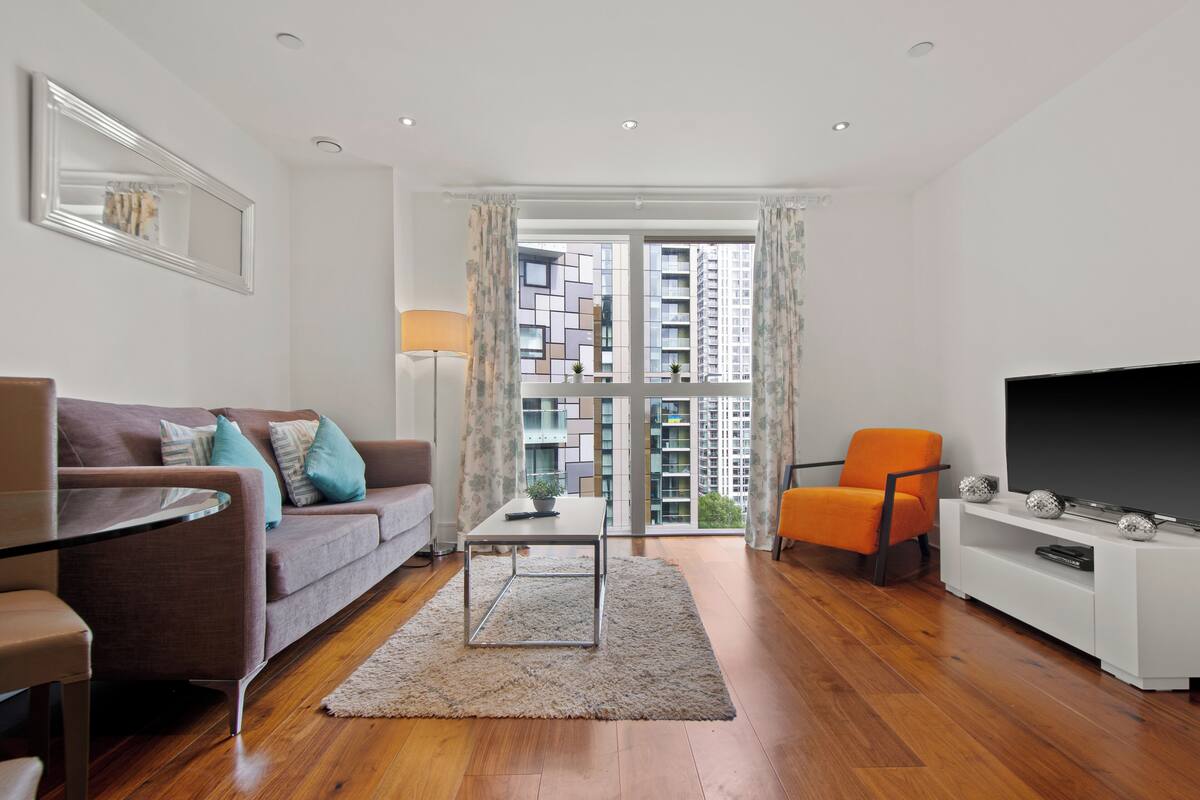 Un salon moderne avec un canapé, une table basse, un téléviseur et une vue sur des immeubles de grande hauteur, visible à travers de grandes fenêtres.