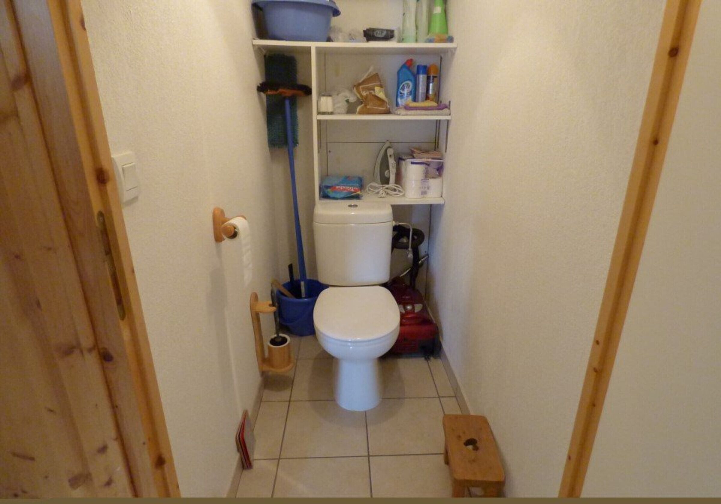 Une petite salle de bain étroite, avec des toilettes blanches, une étagère au-dessus contenant des produits de nettoyage et une porte en bois.