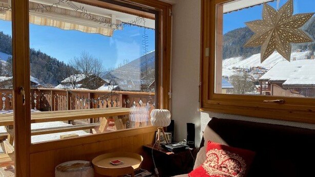 A cozy room with a wooden interior, a window with a view of a snowy mountain landscape, a wooden table with a lamp, and a red pillow on the bed. - Mountain-Style Apartment for 6-7 Near Cable Cars and Town Center in Le Grand-Bornand (Le Grand-Bornand)