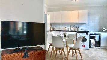 A modern kitchen with white cabinets, a dining area with white chairs, a TV, and a wooden chest of drawers.
