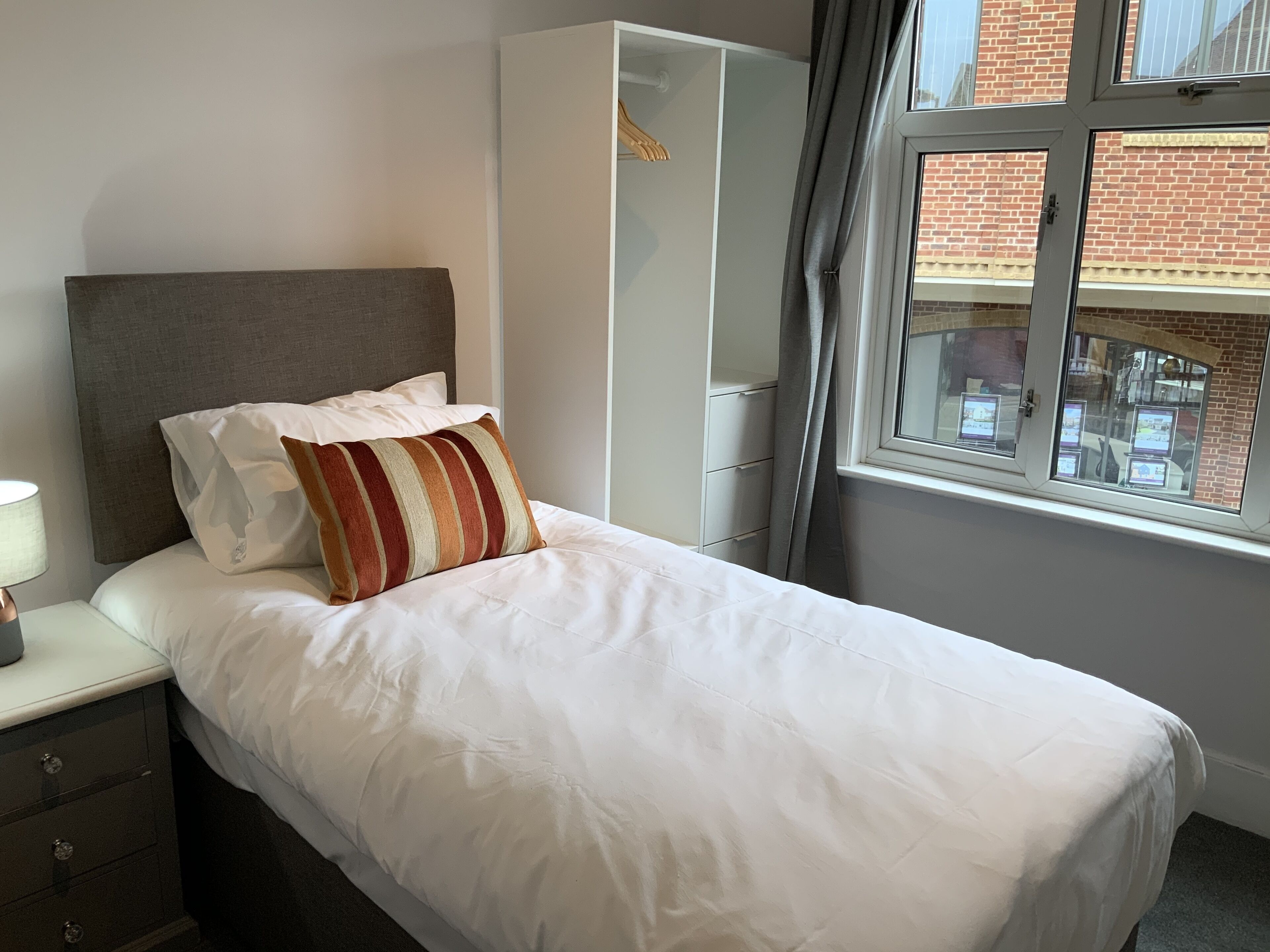 A neatly made bed with white bedding and a striped pillow, a bedside table with a lamp, a wardrobe, and a window with a view of a brick building.