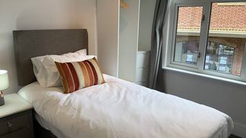 A neatly made bed with white bedding and a striped pillow, a bedside table with a lamp, a wardrobe, and a window with a view of a brick building.