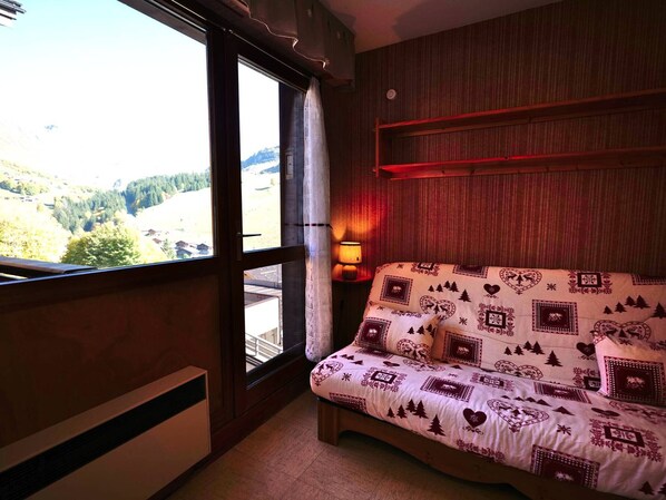 A bedroom with a large window, a bed with patterned bedding, a wall-mounted shelf, and a bedside lamp.