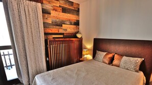 Une chambre à coucher avec une tête de lit en bois, un lit recouvert d’une couverture beige, une table de chevet avec une lampe, et une vue sur l’extérieur par une fenêtre ornée de rideaux.