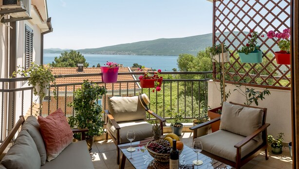 A balcony with a view of a lake, furnished with a sofa, chairs, and a table with a wine bottle and fruit. - Cicciobello Lux Apartment with stunning sea view (Baošići)
