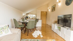 A modern living room with a sofa, a dining area with chairs, a kitchen in the background, a TV on a stand, and a clock on the wall. - Cicciobello Lux Apartment with stunning sea view (Baošići)