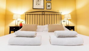 A double bed with two white towels, a headboard with vertical slats, two bedside tables with lamps, and framed pictures on the wall.