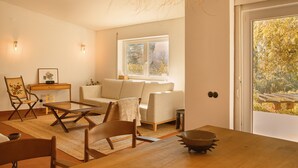 A modern living room with a wooden dining table, a sofa, and a chair. There is a window with a view of trees and a bench outside. - Perfect for quiet vacation with family and friends (Setúbal)