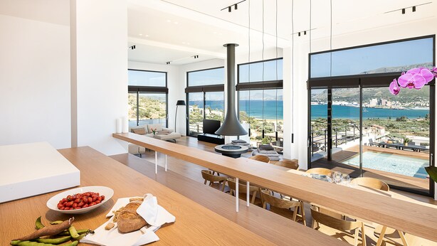Une cuisine moderne avec un long comptoir en bois, un saladier rempli de tomates et une vue sur la mer grâce à de grandes fenêtres.