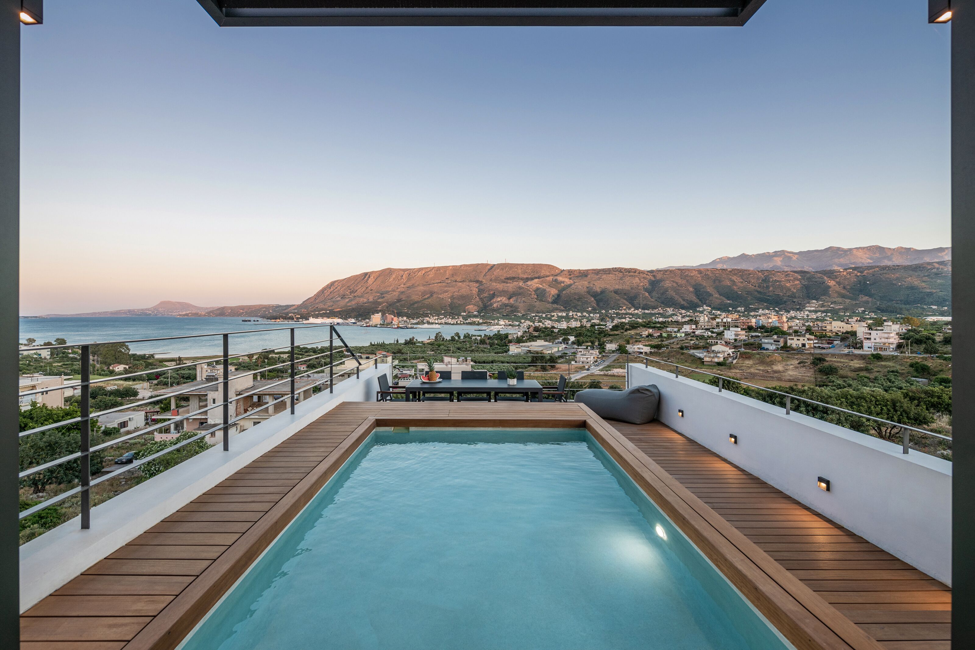 A rooftop pool with a wooden deck, a table with chairs, and a view of a coastal town and mountains.