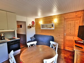 A compact kitchen with modern appliances, a dining area with a wooden table and white chairs, and a cozy seating area with a sofa and cushions.