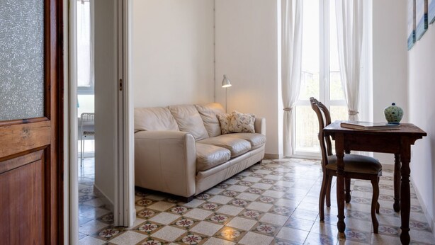 Ein Zimmer mit einem beigefarbenen Sofa, einem Holztisch mit Vase und Buch, einer Lampe und einem gemusterten Boden.