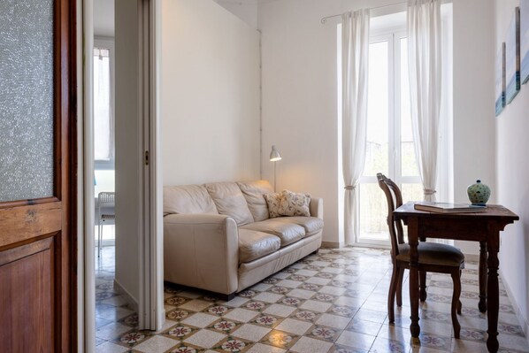 Una habitación con un sofá color crema, una mesa de madera con un jarrón y un libro, una lámpara y un suelo estampado.