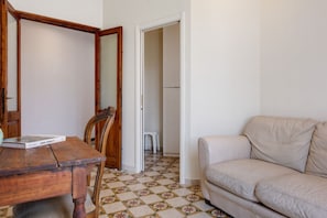 Ein Zimmer mit einem Holztisch, einem beige farbenen Sofa, einem Fliesenboden und einer Tür zu einem weiteren Raum.