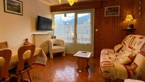 Una sala de estar acogedora con suelo de madera, un sofá, una mesa de comedor con sillas, un televisor y una ventana con vistas a las montañas.