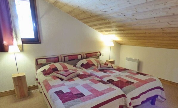 Een slaapkamer met twee bedden, houten balken in het plafond, een raam met gordijnen en een bedlampje.