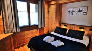 A wooden bedroom with a bed, two pillows, a nightstand, a window with curtains, and a wall-mounted artwork. - Charming 3-room renovated with sauna and jacuzzi for 6 in Grand-Bornand (Le Grand-Bornand)