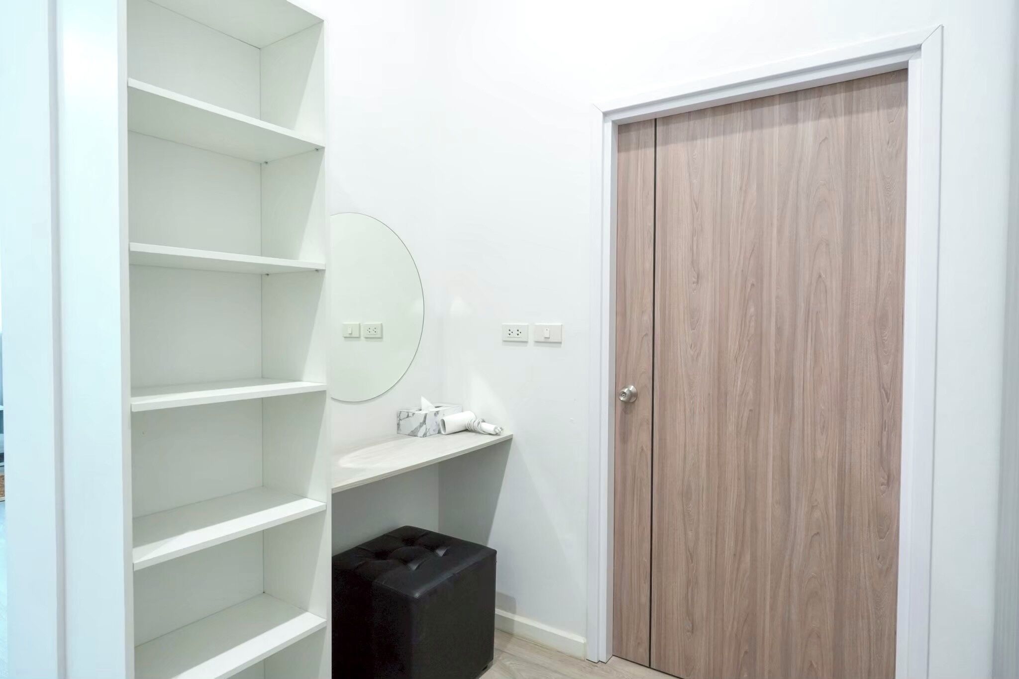 Una habitación con paredes blancas, una puerta de madera, un espejo y un caja de almacenamiento negra.