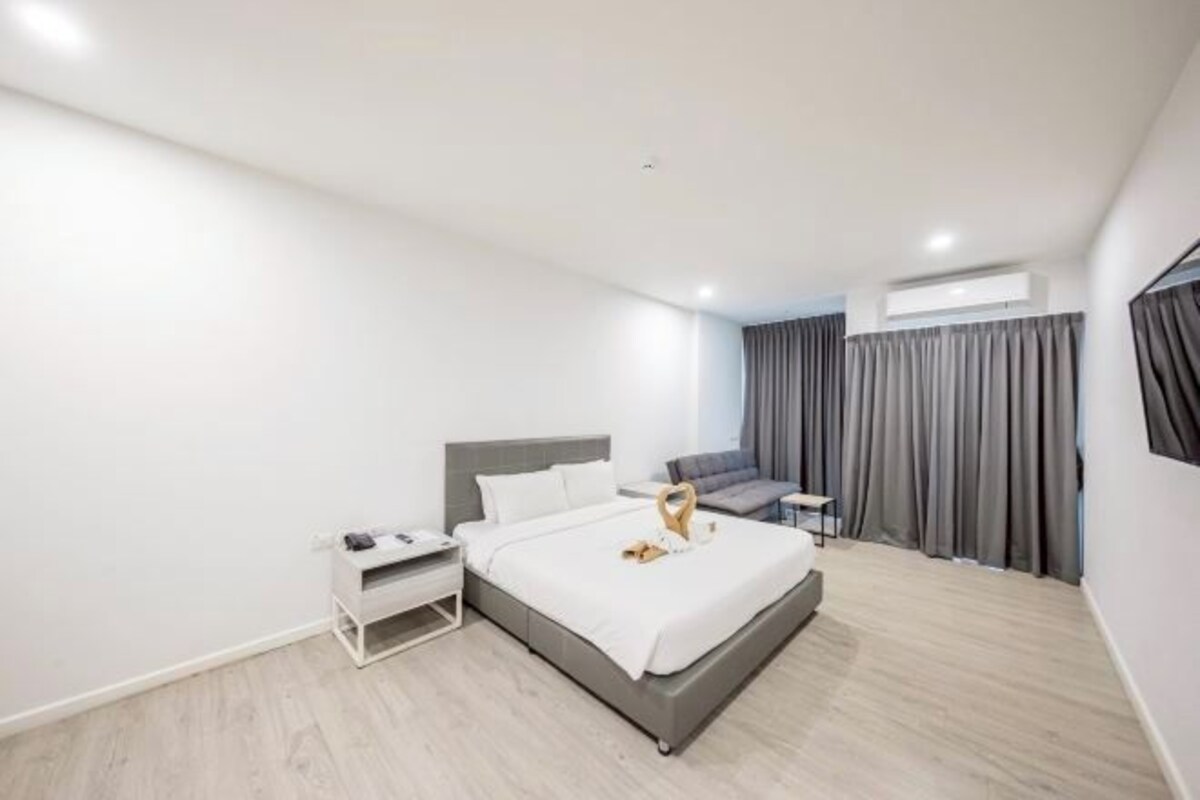 Habitación de hotel moderna con cama, mesita de noche y zona de estar con sofá y cortinas.