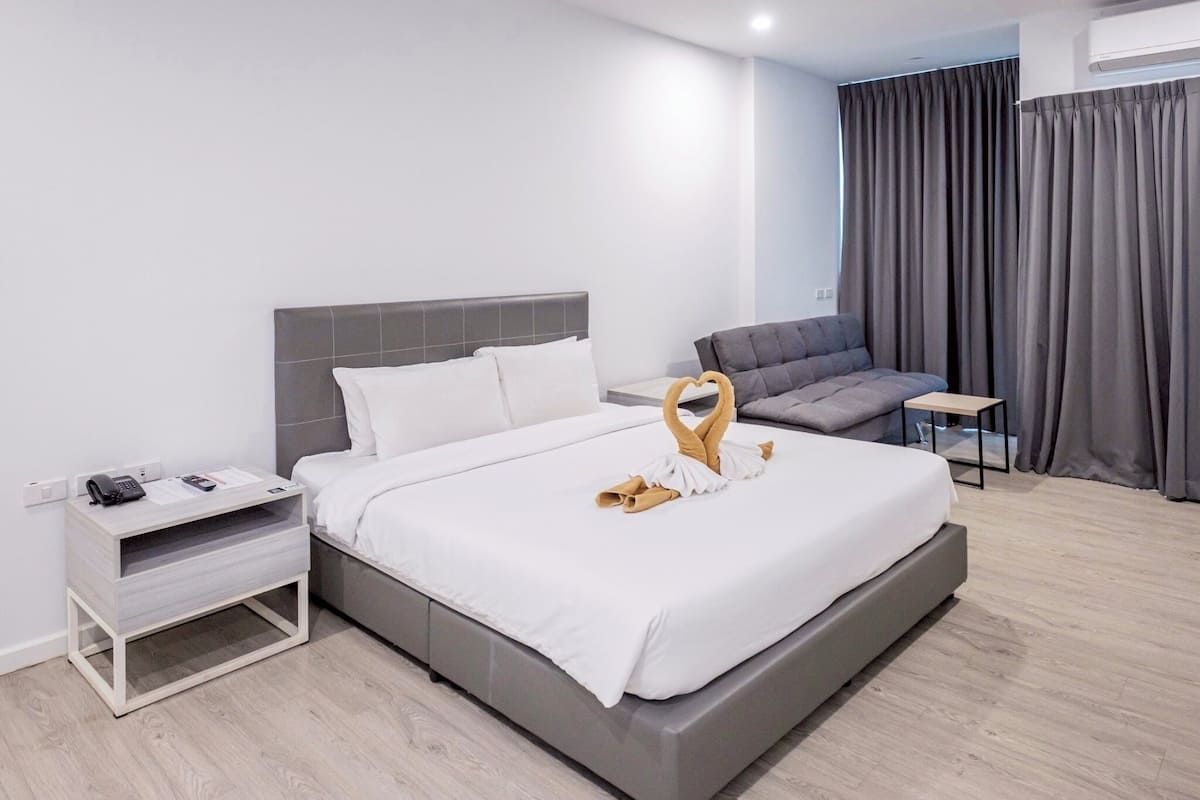 A modern hotel room with a large bed, a bedside table, a small desk, a sofa, and a window with curtains.