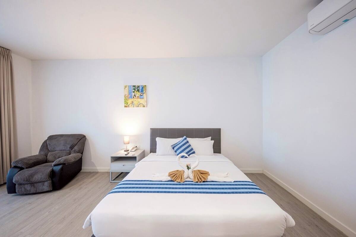 A hotel room with a bed, a chair, a nightstand, and a wall-mounted air conditioner.