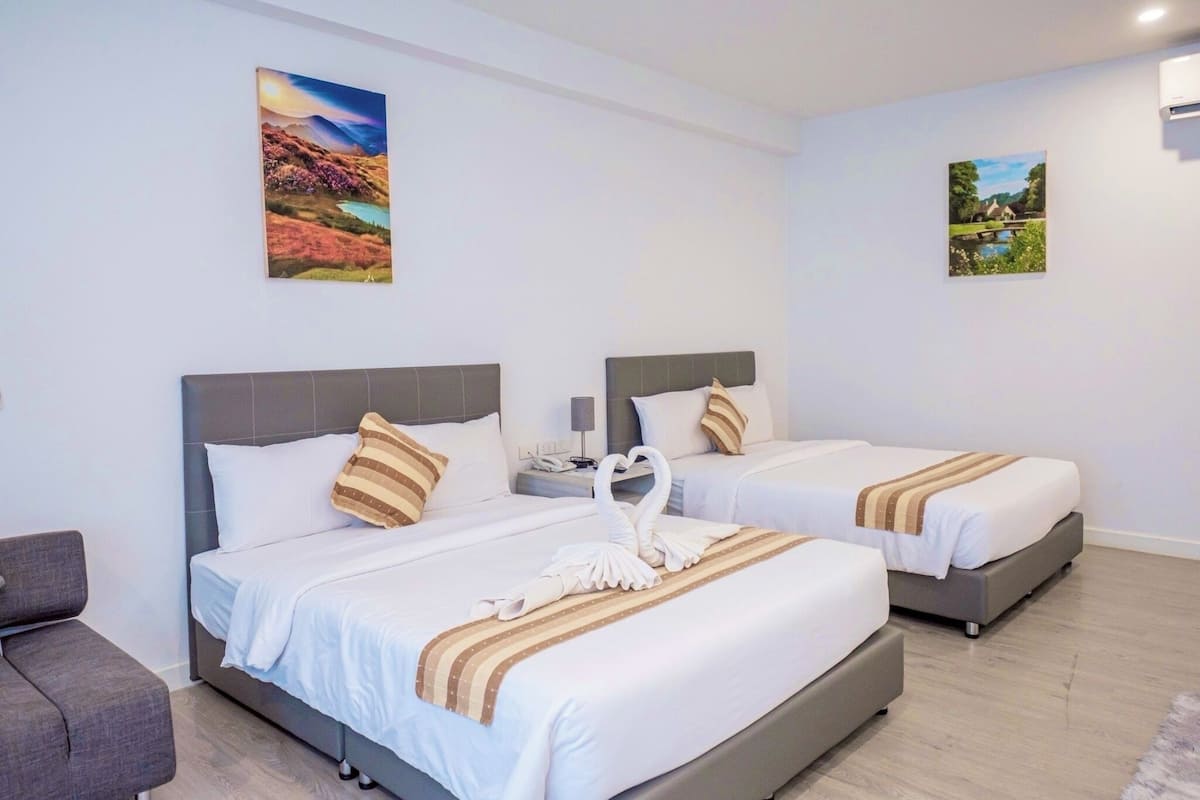 A hotel room with two beds, each with a headboard, pillows, and a blanket. There is a nightstand with a lamp and a phone. The room has a light-colored floor and white walls.