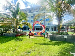 Entrada de un hotel con palmeras, un cartel que dice @GrandPA y una piscina al fondo.