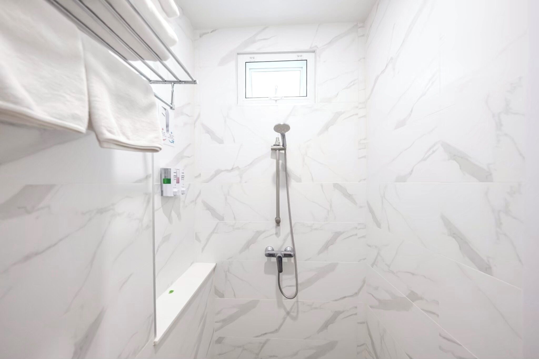 A bathroom with a shower, a towel rack, a window, and a white shelf.