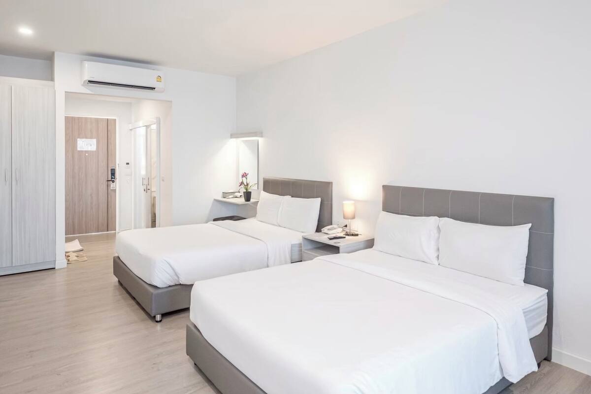 A hotel room with two beds, a bedside table with a lamp, an air conditioning unit, and a door leading to another room.