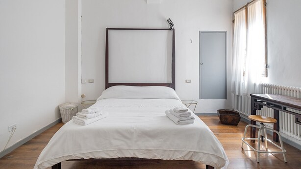 A minimalist bedroom with a large bed, a wooden floor, a radiator, and a small stool. - A pochi passi da Piazza Maggiore by Wonderful Italy (Bologna)