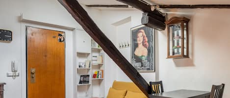 A room with a wooden door, a yellow sofa, a dining table with chairs, a bookshelf, and a poster on the wall.
