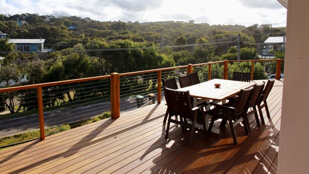 A wooden deck with a table and chairs, overlooking a road and greenery.