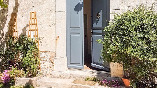 Un bâtiment en pierre avec une porte grise, un petit jardin agrémenté de plantes en pot et une échelle en bois appuyée contre le mur.