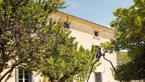 Un puits en pierre avec un robinet en métal, situé devant un bâtiment doté d’un toit en tuiles et d’une fenêtre avec des volets.