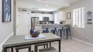 Una cocina moderna con armarios blancos, un refrigerador de acero inoxidable y un microondas. Hay un comedor con una mesa de madera y sillas azules.