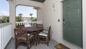 Un porche techado con una mesa y sillas de madera, una puerta verde y vistas a palmeras y un edificio al fondo.