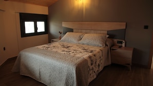 Une chambre avec un grand lit, une tête de lit en bois, des tables de chevet, une lampe fixée au mur et une fenêtre avec des stores.