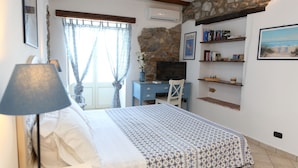 A bedroom with a bed, a desk, a TV, and a stone wall. - Room with balcony and beautiful sea view in the old town (Cefalù)