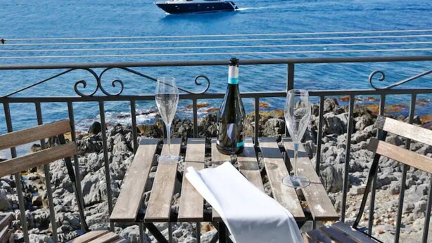 A wooden table with a bottle and glasses, overlooking a rocky shoreline and a yacht on the water. - Room with balcony and beautiful sea view in the old town (Cefalù)