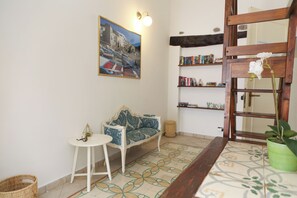 A cozy living area with a patterned sofa, a small round table, a bookshelf, and a decorative wall painting. - Suite 58 · Two Ensuite Bedrooms, Sea & Old-Town Balconies, 1 Min to the Beach (Cefalù)