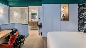 A modern hotel room with a bed, a desk, a chair, a wardrobe, and a bathroom visible through an open door.