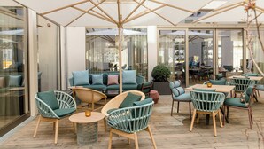 An outdoor seating area with wicker chairs and a wooden table, a sofa with cushions, and a large umbrella. - Motel One Aachen (Aachen)