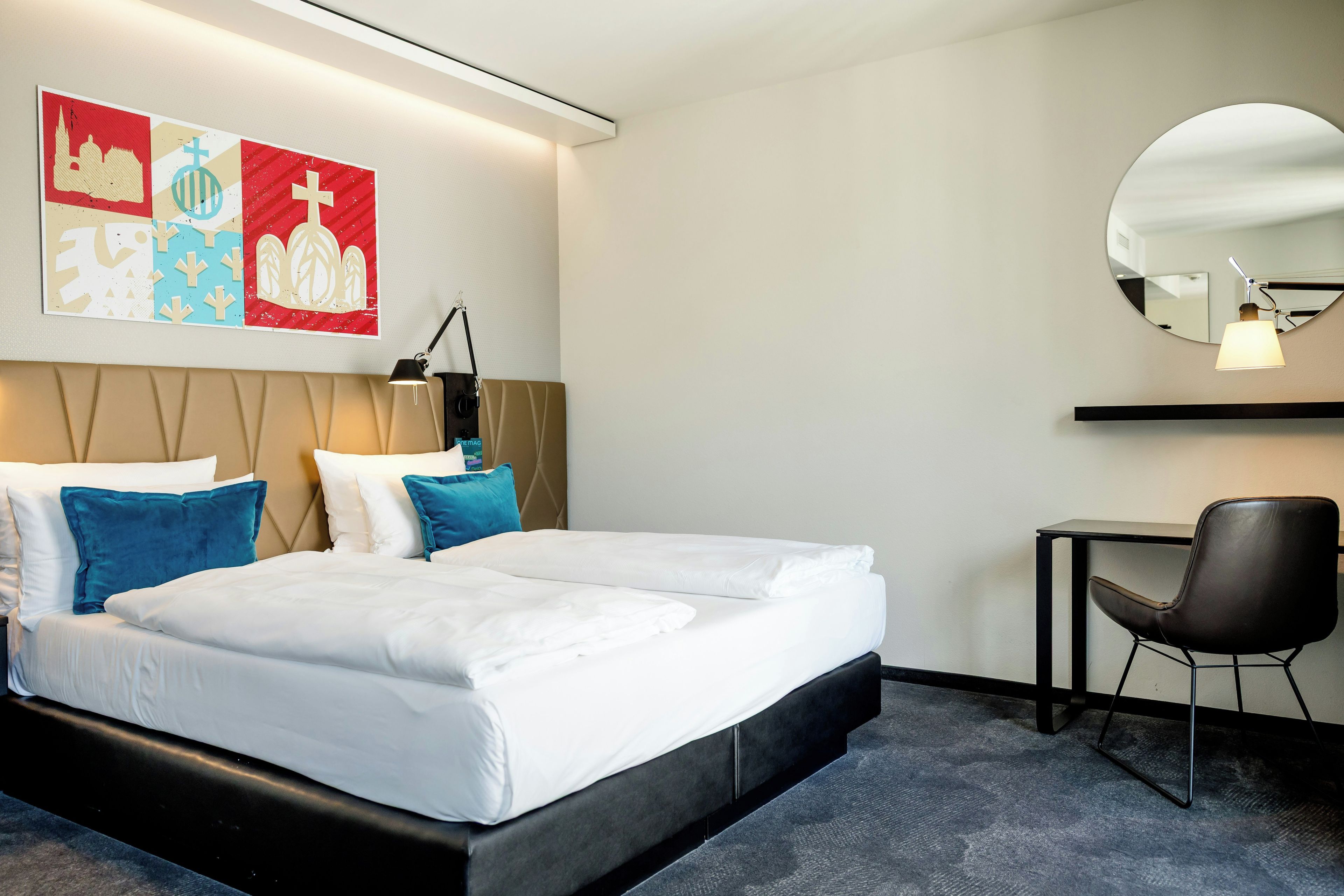 A hotel room with a bed, pillows, a desk, a chair, and wall art.