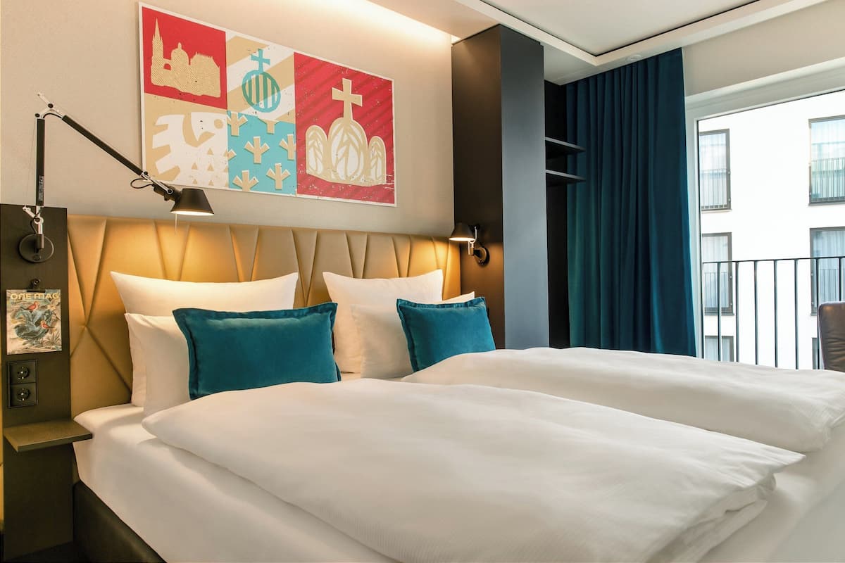 Ein Hotelzimmer mit einem großen Bett, einem Kopfteil mit einer Leuchte, einem Wandbild, einem Nachttisch mit einer Lampe und Blick ins Freie durch ein Vorhangfenster.