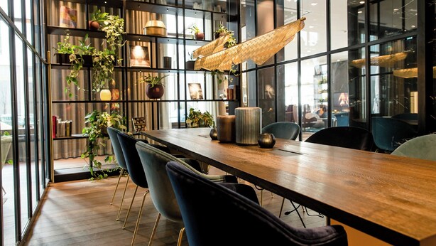 A modern conference room with a long wooden table, black chairs, and a bookshelf filled with plants and books. - Motel One Aachen (Aachen)